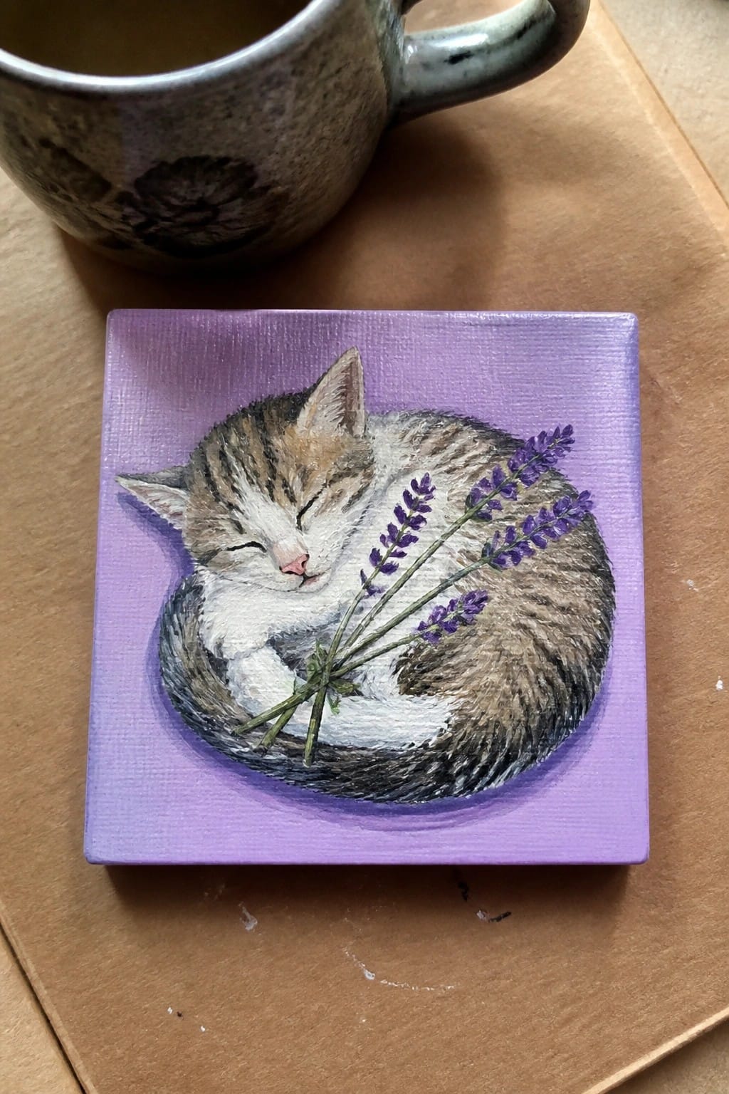 Mini canvas painting of sleeping tabby kitten with lavender on purple background beside a mug.