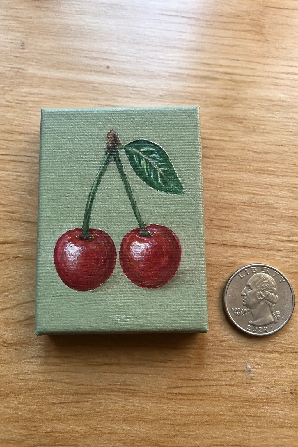 Miniature painting of two red cherries on green canvas beside a 2022 quarter
