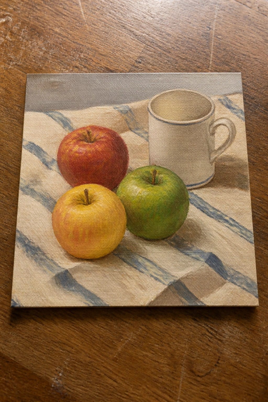 Still life painting of three apples and white mug on blue-striped cloth