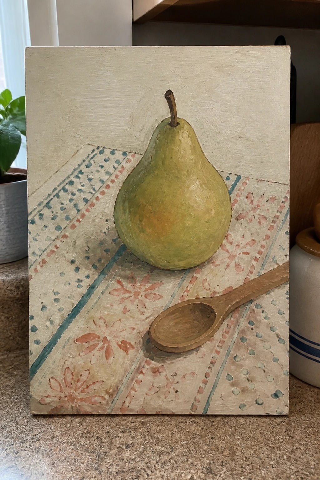 Green pear with stem beside wooden spoon on floral patterned cloth painting