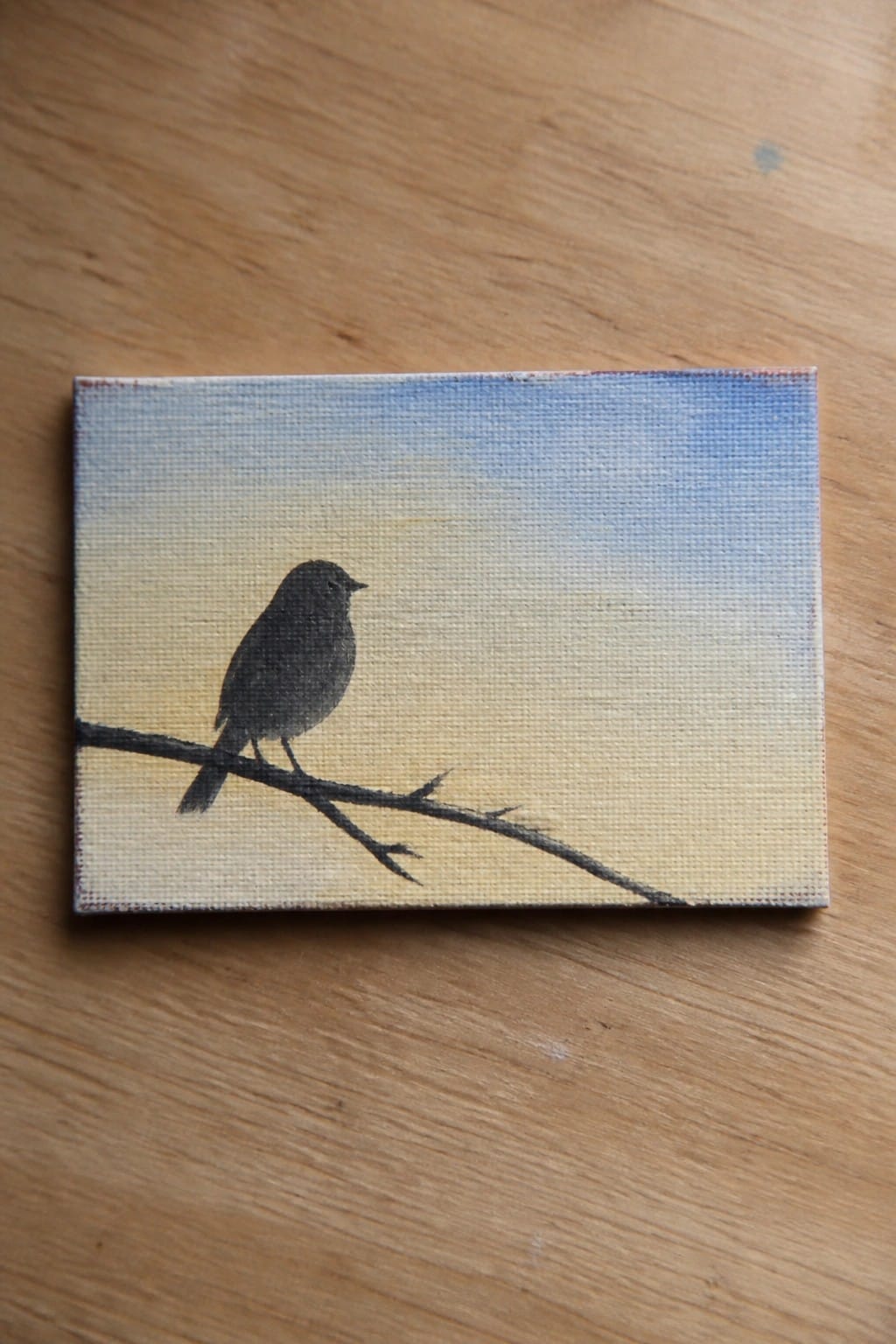 Miniature canvas painting of black bird silhouette on branch against blue-yellow sky gradient on wood.