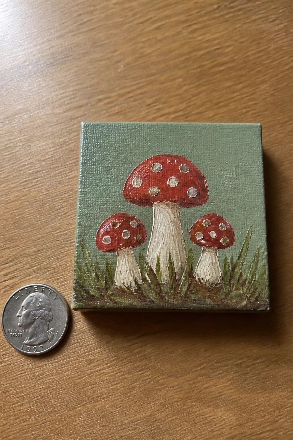 Tiny painting of three red spotted mushrooms beside a 1997 quarter coin