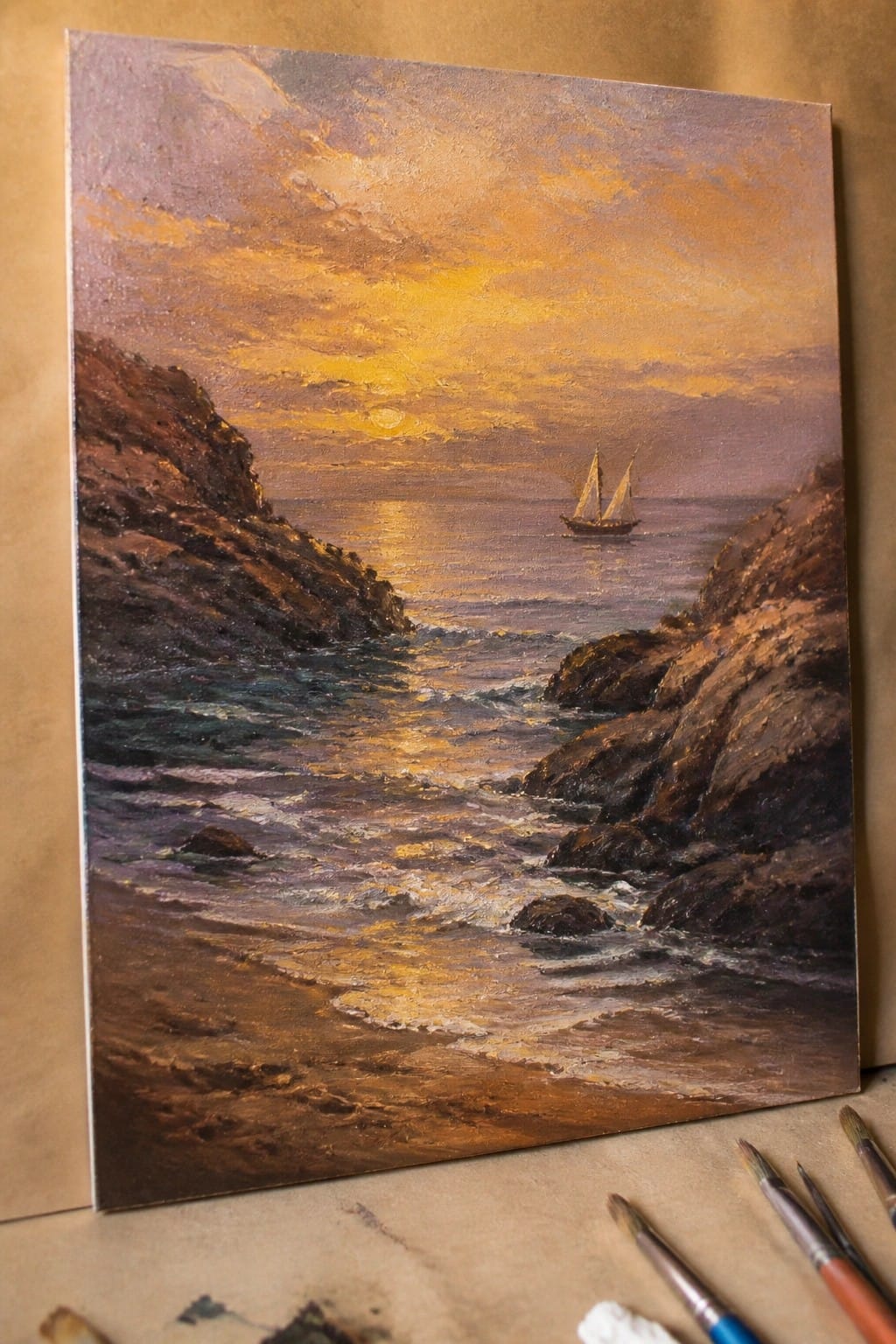 Seascape oil painting of sunset with sailboat between rocky cliffs and paintbrushes nearby.