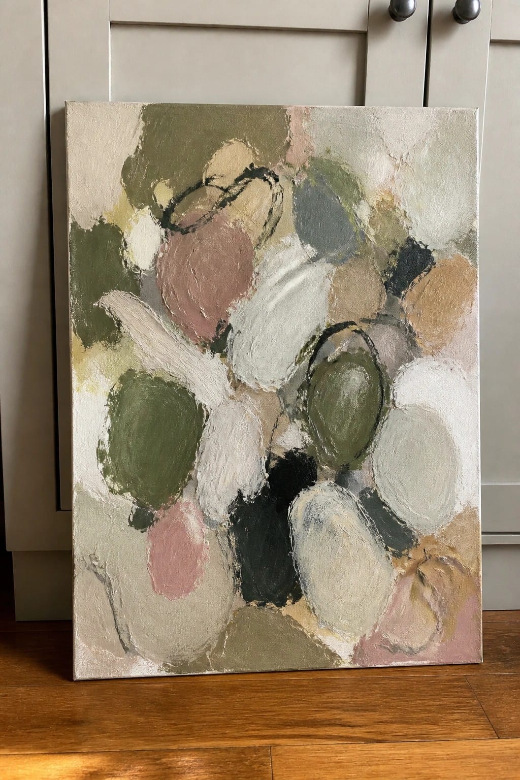 Abstract painting with muted green, pink, and white shapes leaning against cabinet on wood floor