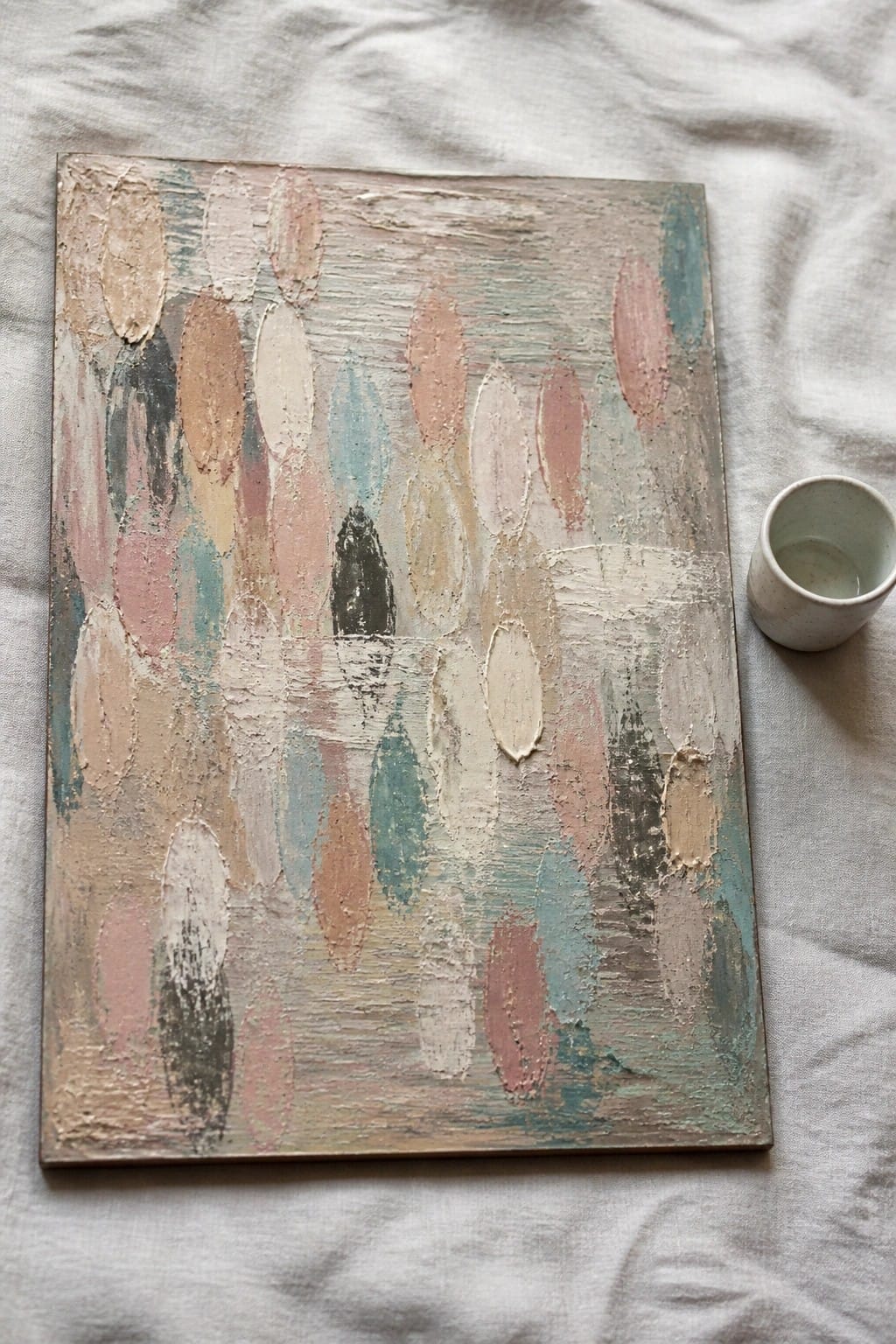An abstract oil painting with textured oval shapes in pastel colors on a canvas next to a small cup.
