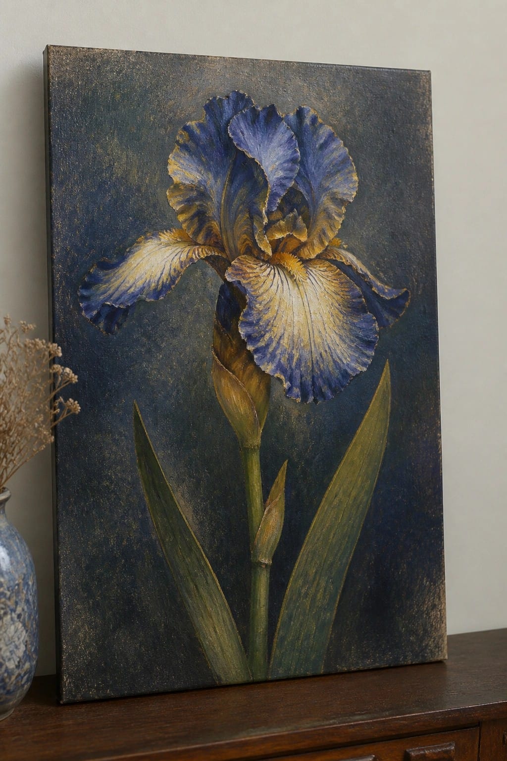 An oil painting of a blue and yellow iris flower on a dark textured background, displayed on a wooden surface next to a blue vase with dried flowers.