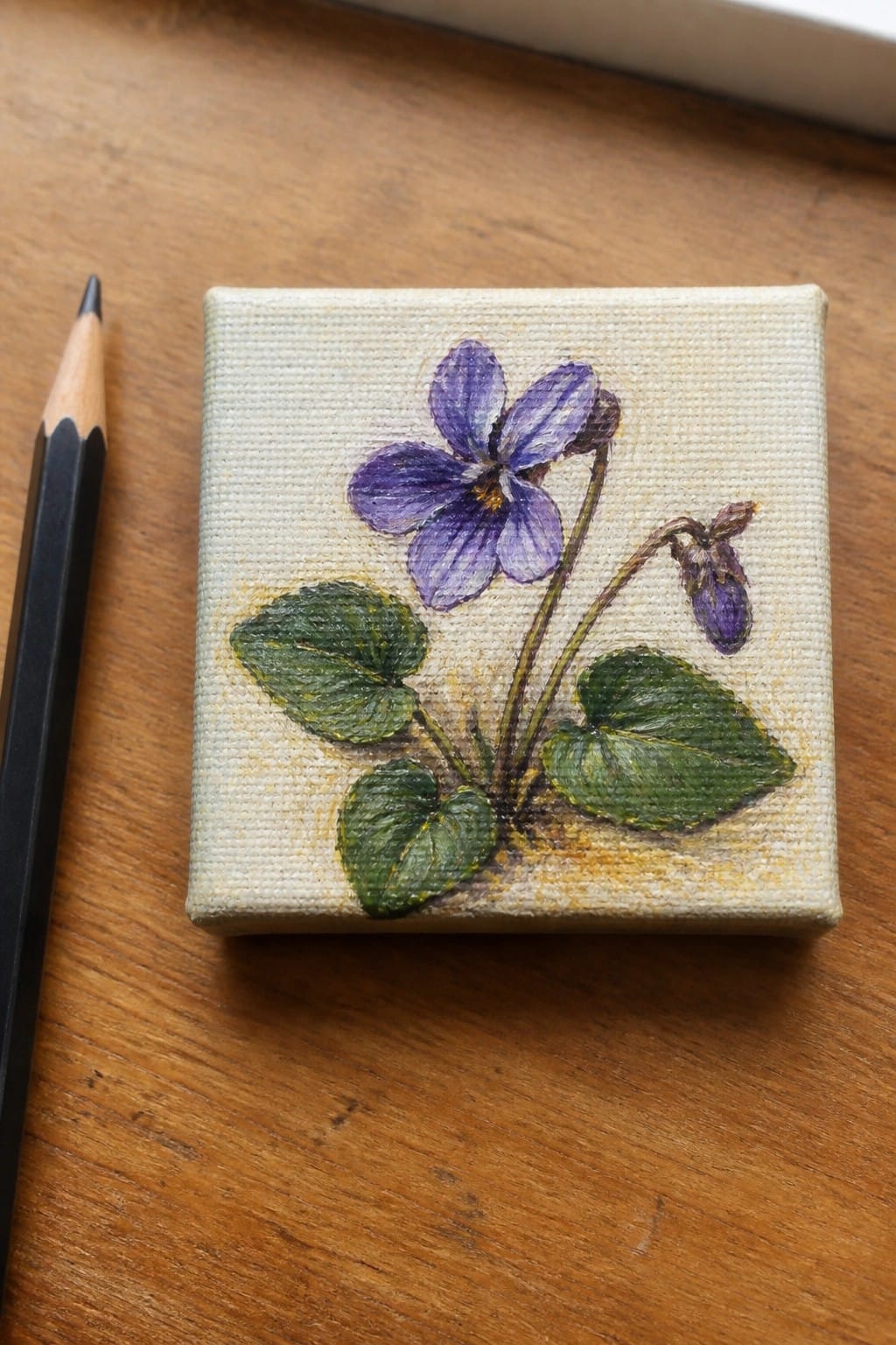 Oil painting of a purple violet with leaves and a bud on a small canvas beside a pencil.