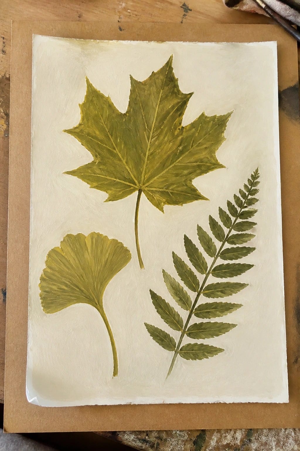 Three green-yellow painted leaves—a maple, ginkgo, and fern—on off-white paper.