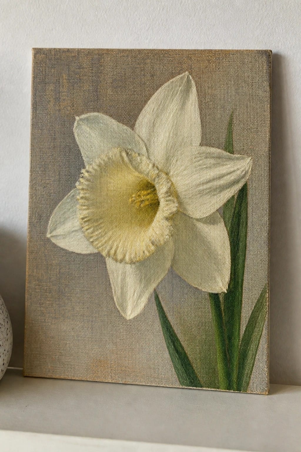 White daffodil with yellow center and green leaves on textured canvas.