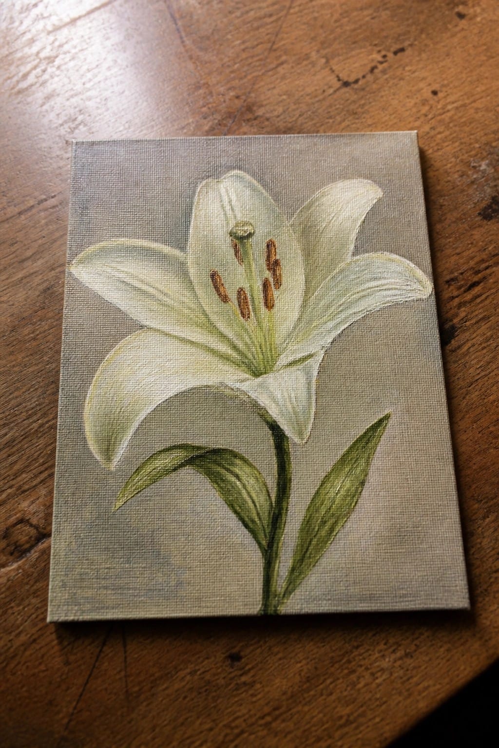 White lily with green leaves painted on canvas atop wooden table