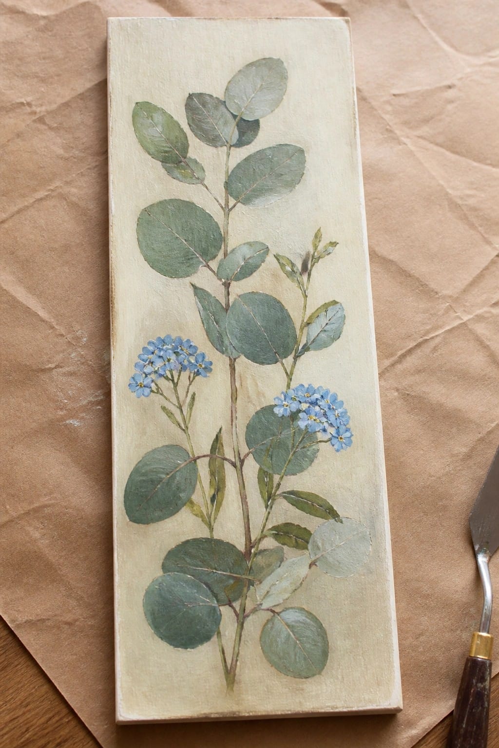 Botanical painting of eucalyptus leaves and blue flowers on a rectangular panel.