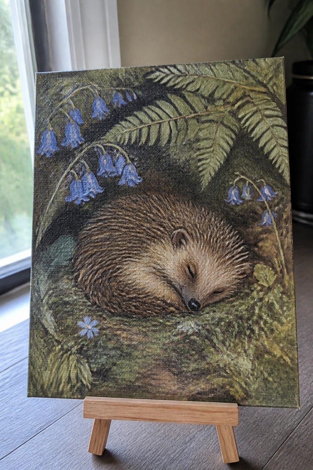 Curled sleeping hedgehog amid bluebells and ferns in realistic painting on easel