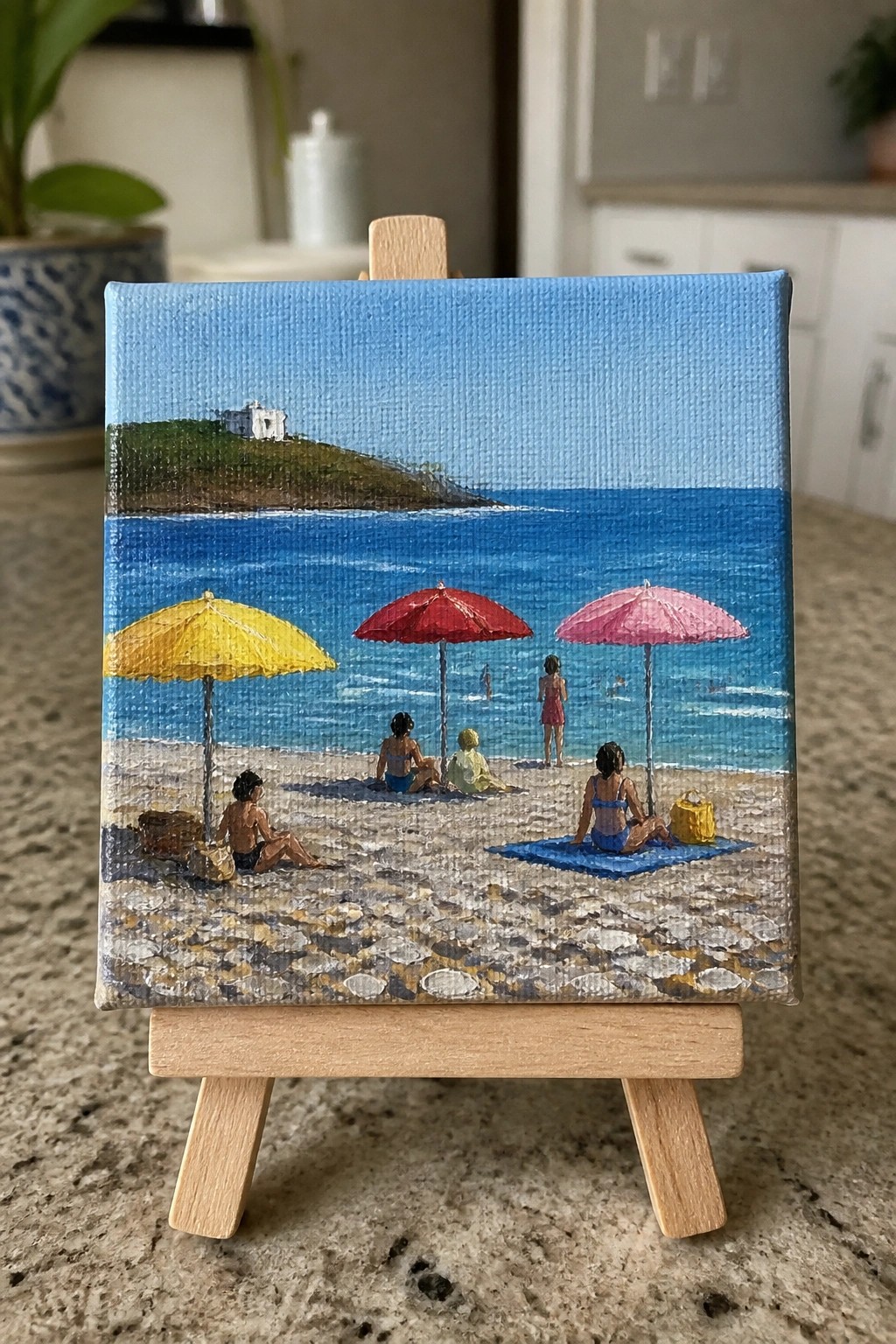 Miniature beach painting on easel with yellow, red, pink umbrellas, ocean, and distant hill.