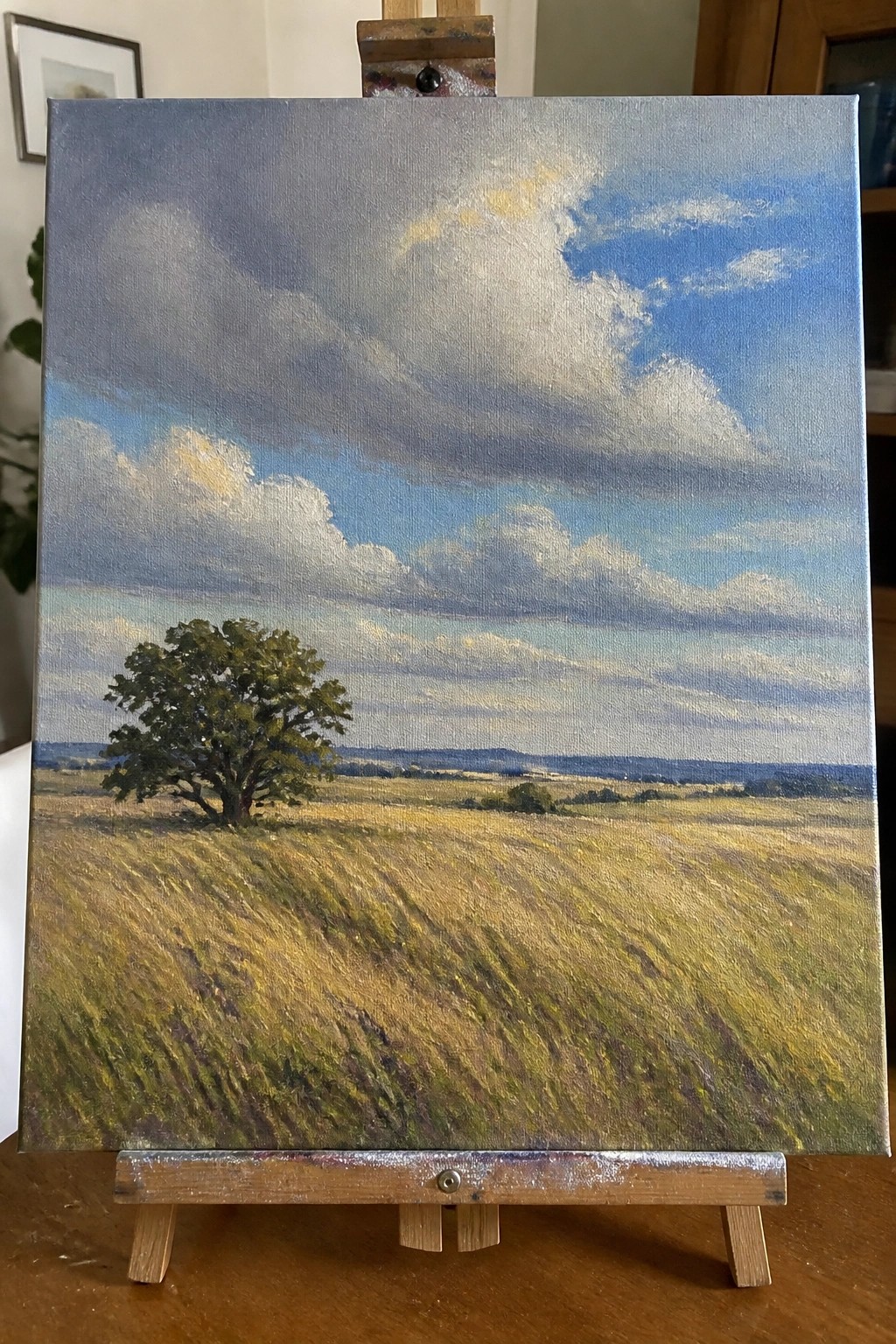 Landscape painting on easel of golden field with lone tree under cloudy blue sky