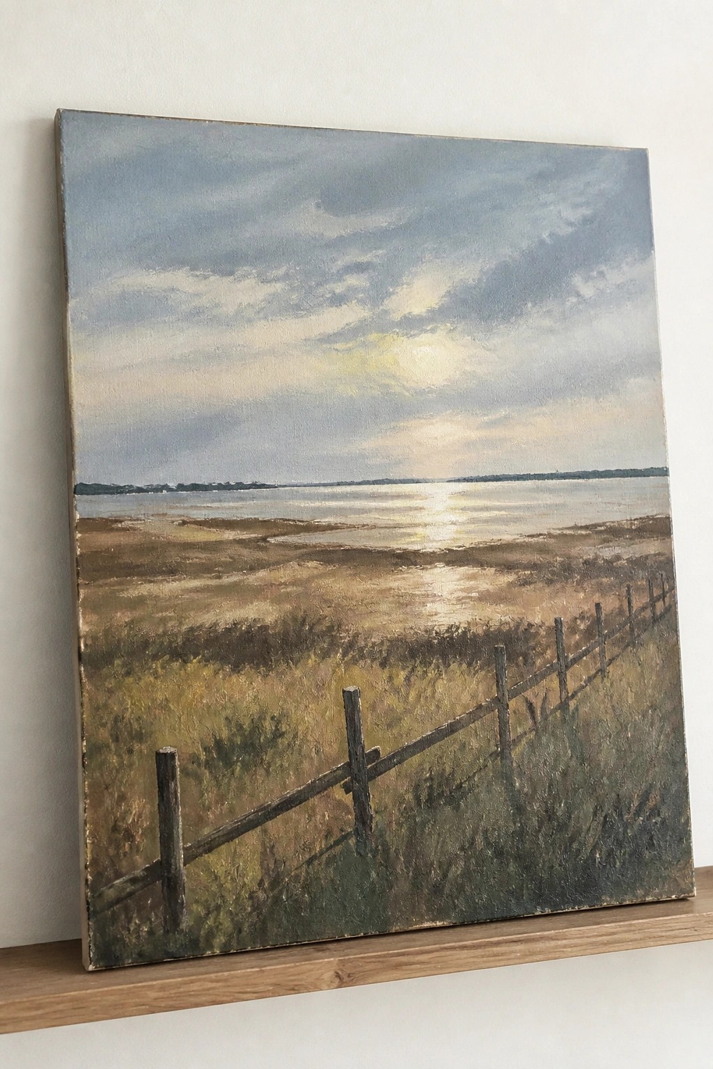 Wooden fence borders marshy field beside sunlit water under cloudy sky.