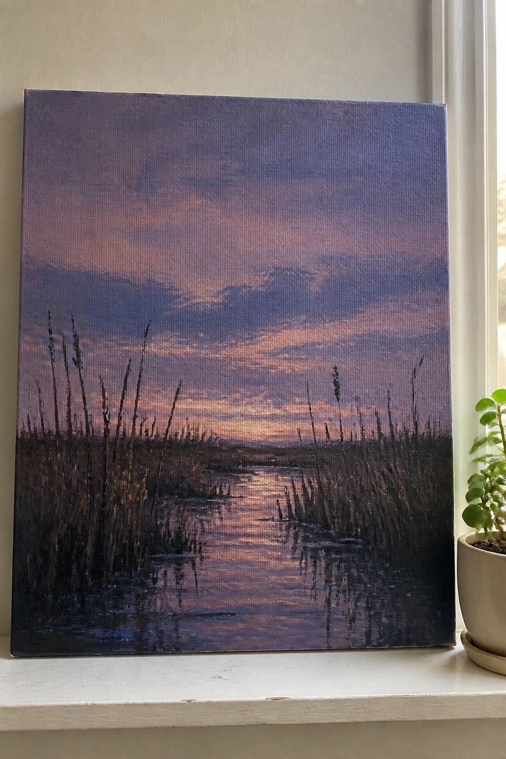 Painting of sunset over reeds and water on windowsill beside potted plant