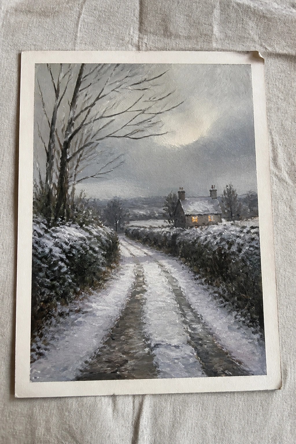 Snowy path with tire tracks leads to lit stone cottage amid winter hedges and trees