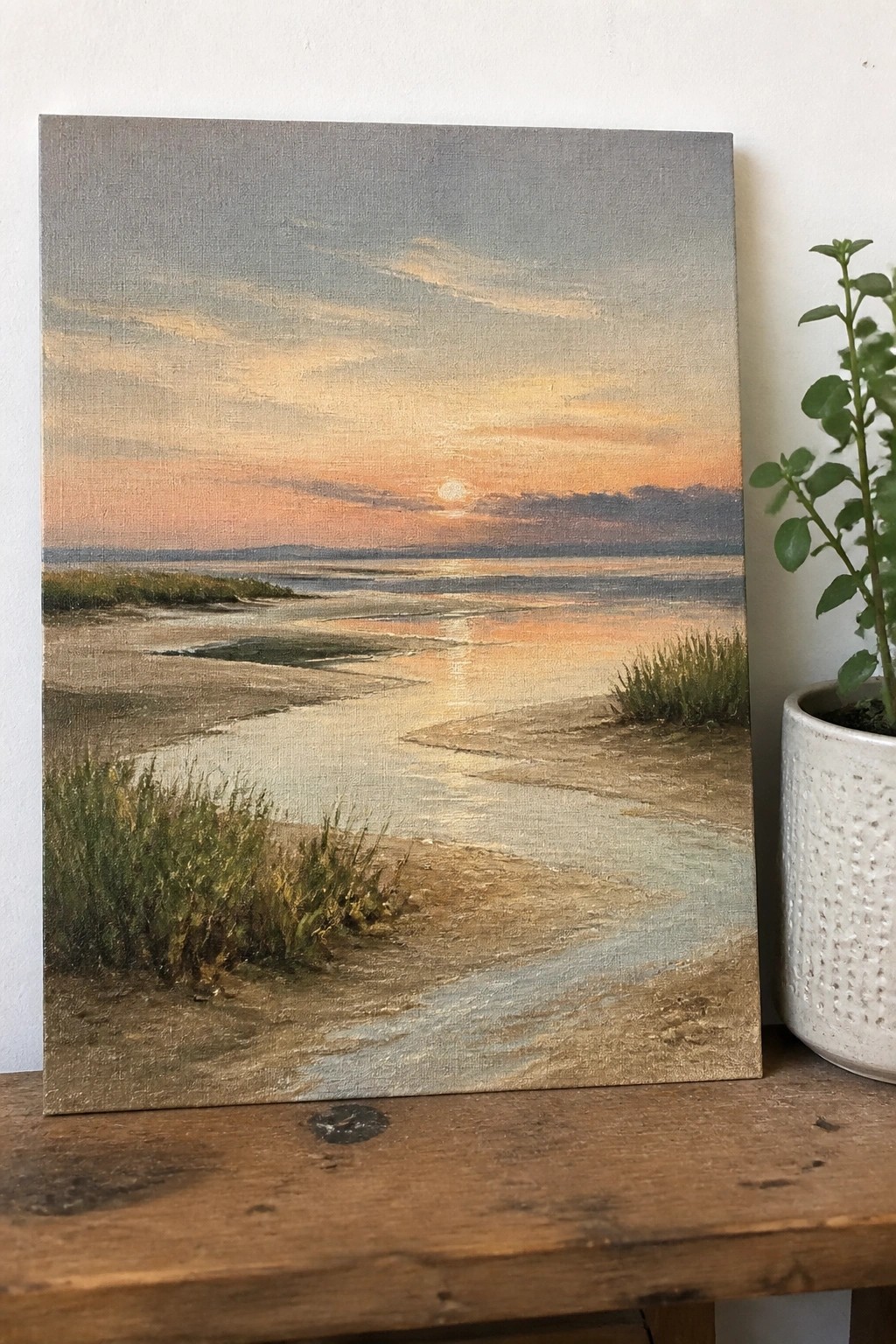 Sunset marsh painting on canvas beside potted plant on wooden table.