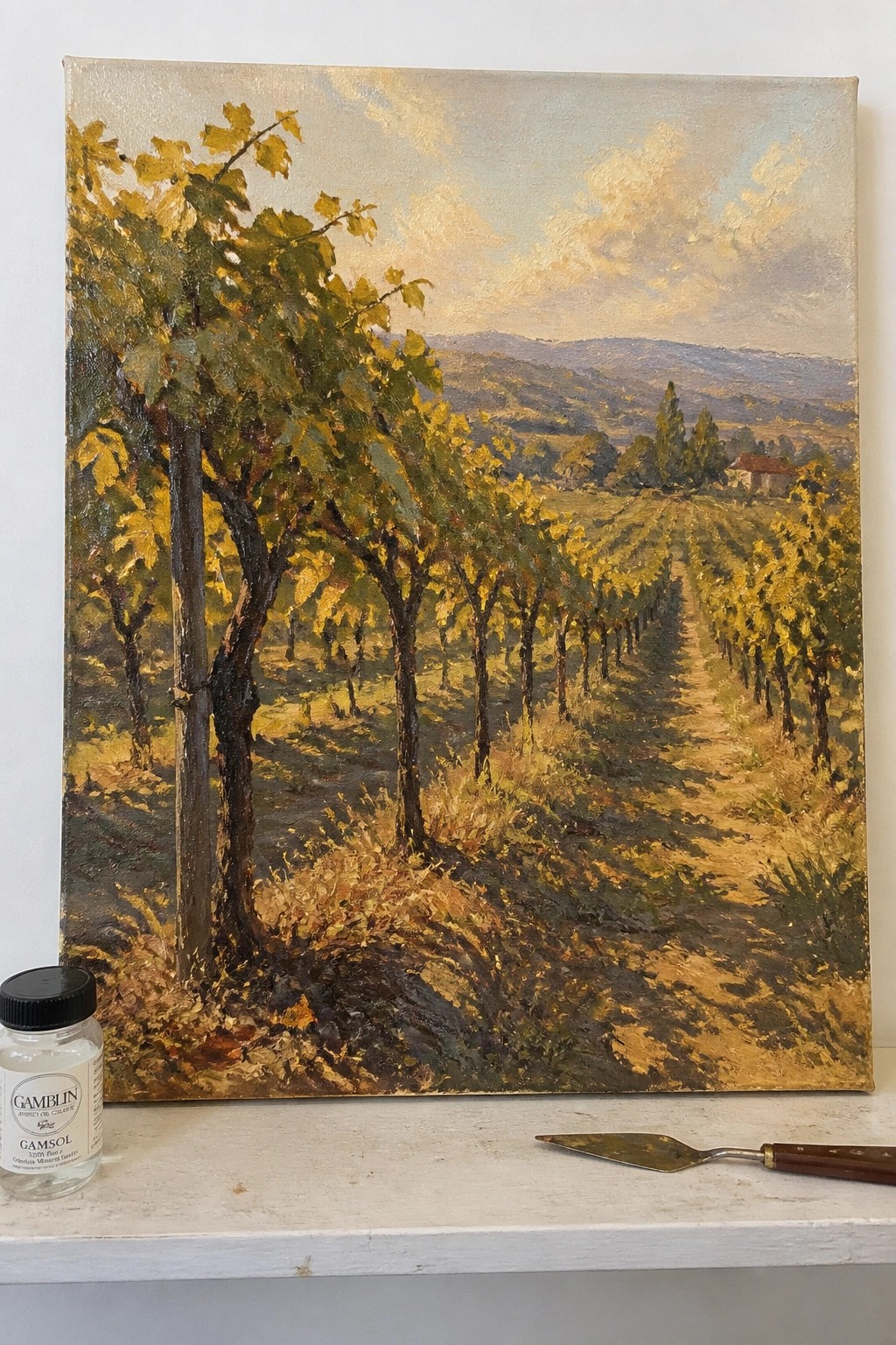 Oil painting of vineyard path with yellow trees, hills, and art supplies on table