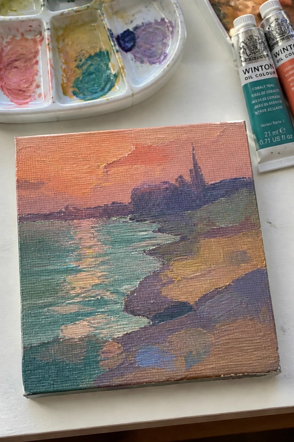 Small canvas with sunset landscape painting beside palette and oil paint tubes
