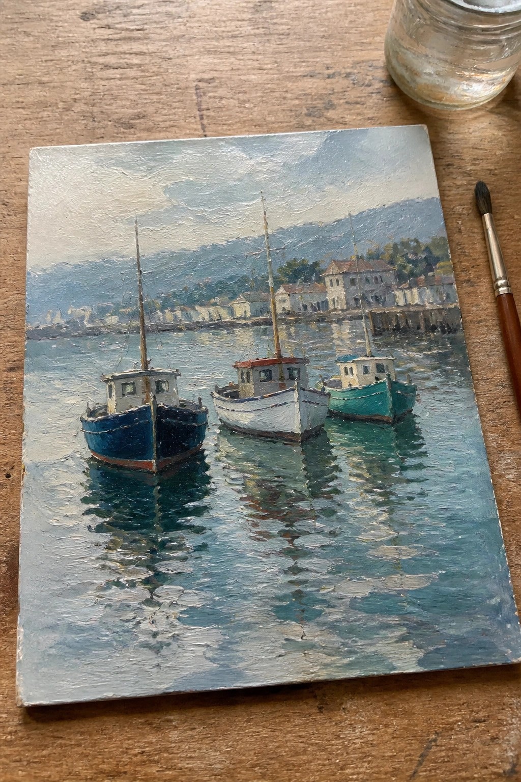 Three boats moored in calm harbor, oil painting with hills and buildings behind, paintbrush nearby.