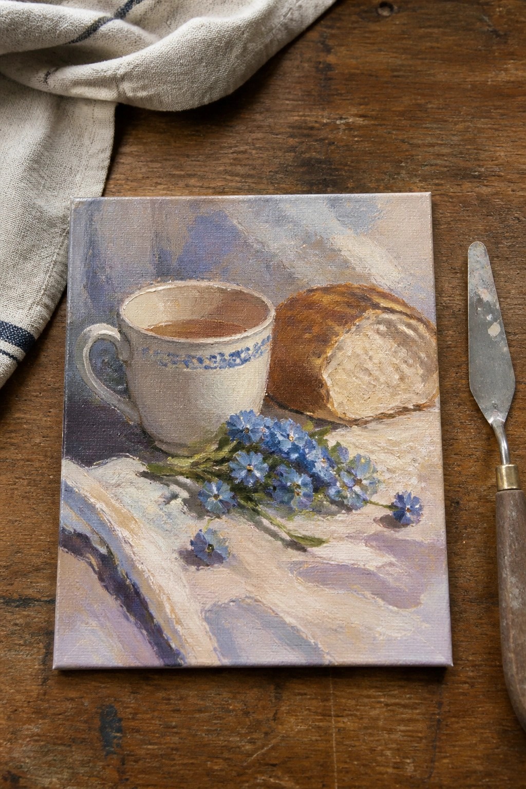 Still life painting of white teacup, bread loaf, blue flowers beside palette knife on wood.