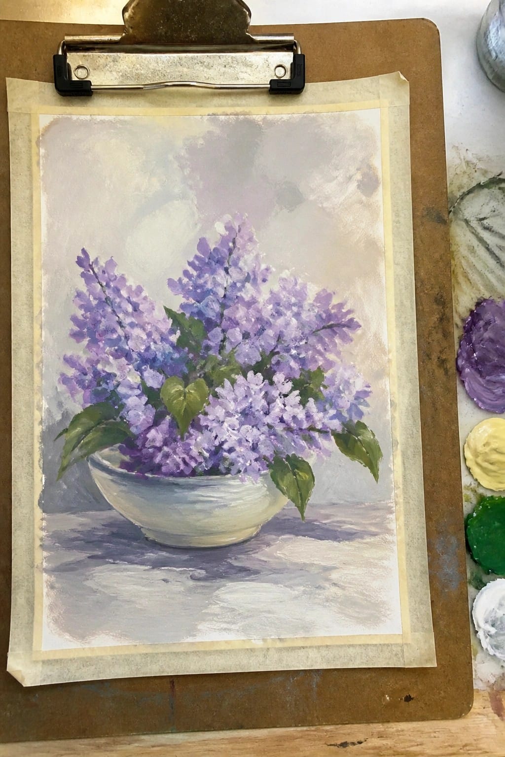 Painting of purple lilacs in white bowl on clipboard beside paint palette