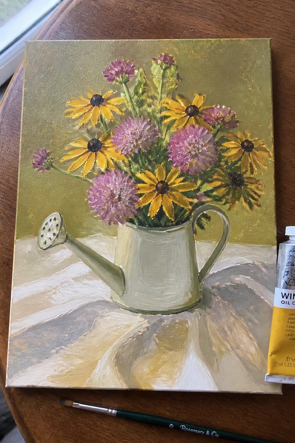 Oil painting of yellow and purple flowers in green watering can with paint tube and brush nearby