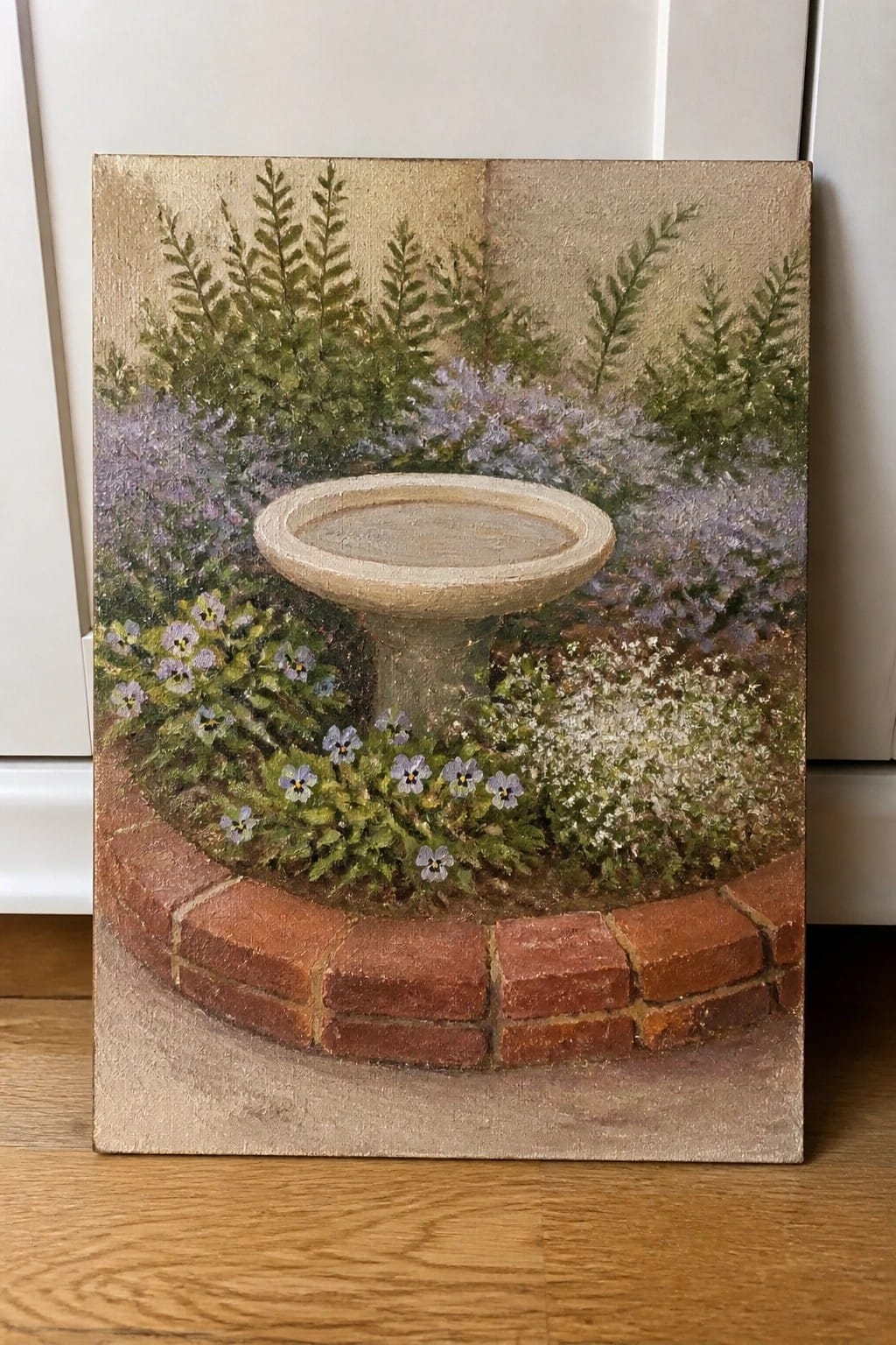 Oil painting of a stone birdbath encircled by bricks amid purple flowers and greenery.