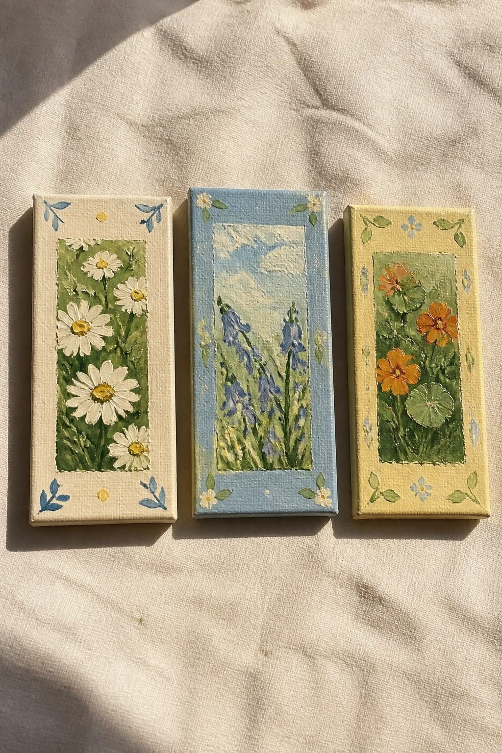 Three small floral paintings on canvas with daisies, bluebells, and orange flowers on fabric.