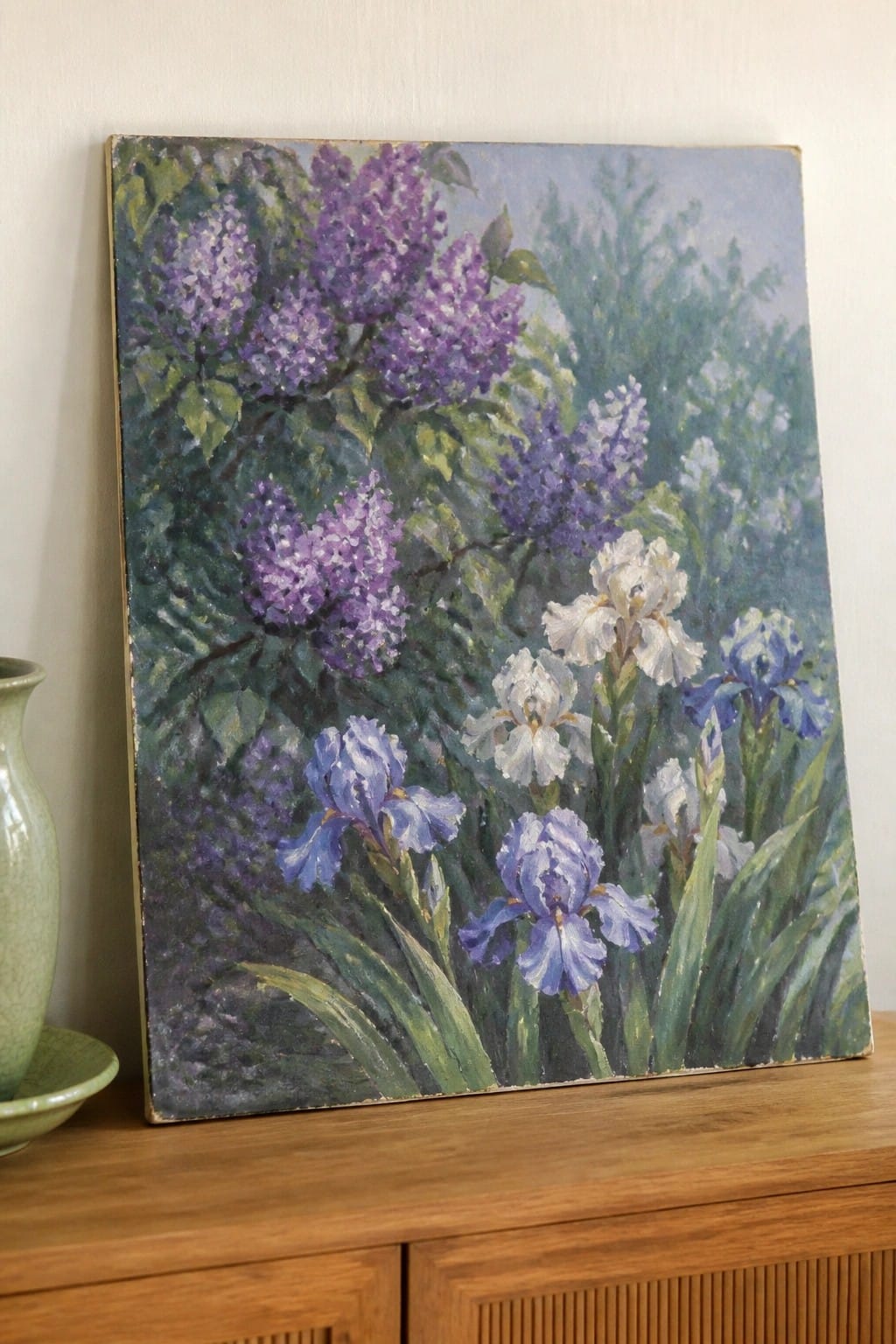 Oil painting of purple lilacs and blue-white irises on wooden cabinet beside vase