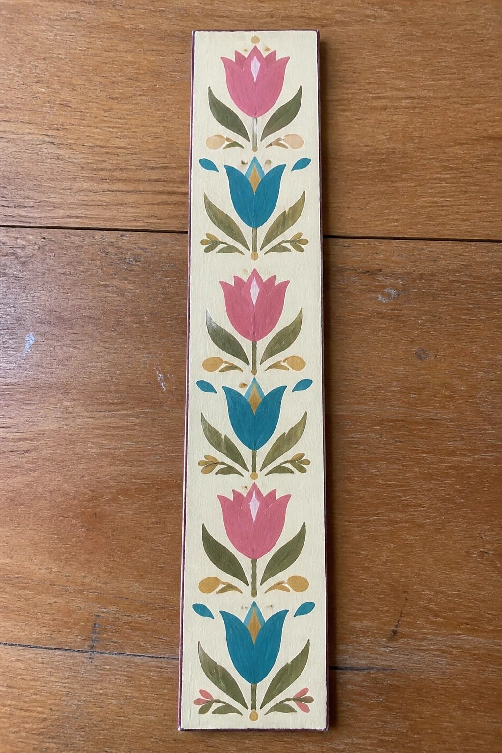 Vertical wooden board with a repeating painted pattern of pink tulips, blue flowers, green leaves, and small accents on a cream background.