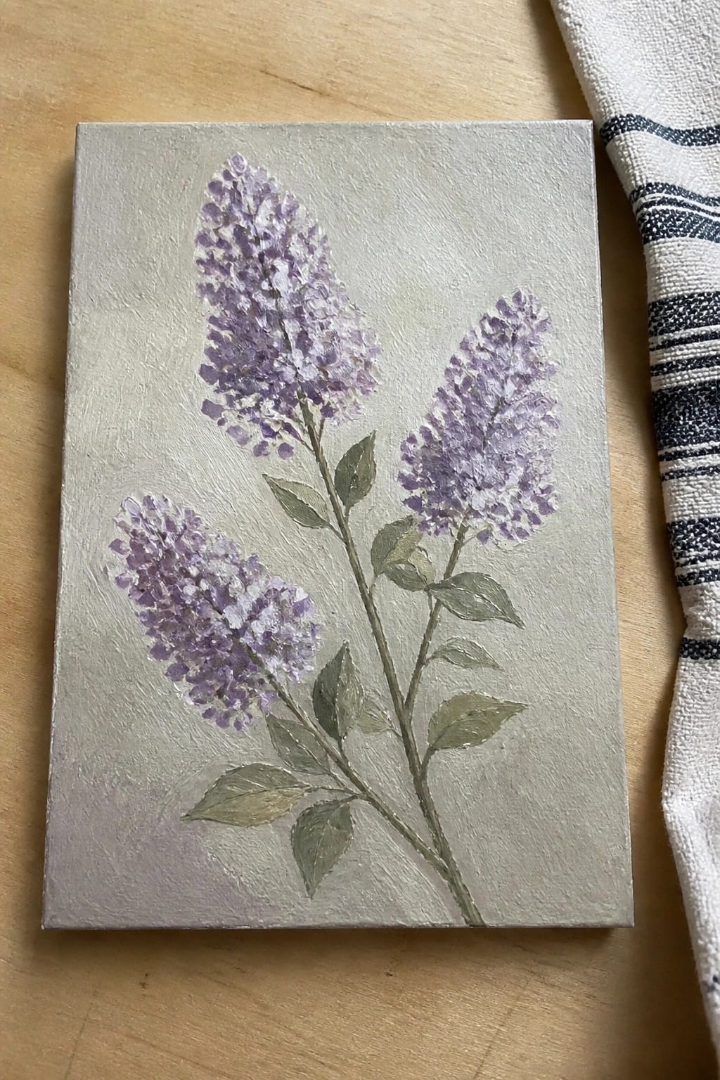 Small canvas with three purple lilac clusters and green leaves on wood surface