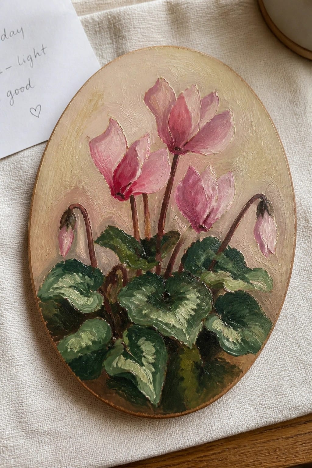 An oval oil painting of pink cyclamen flowers with green leaves on a neutral background.