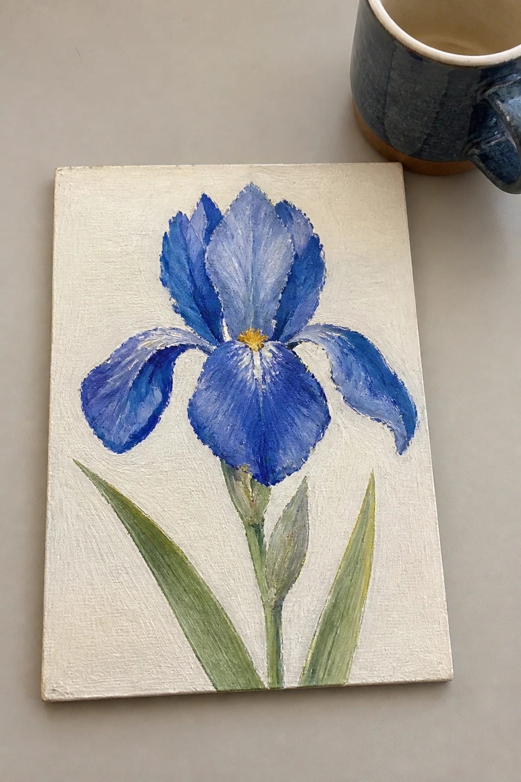 Blue iris painting on white canvas beside blue ceramic mug on table