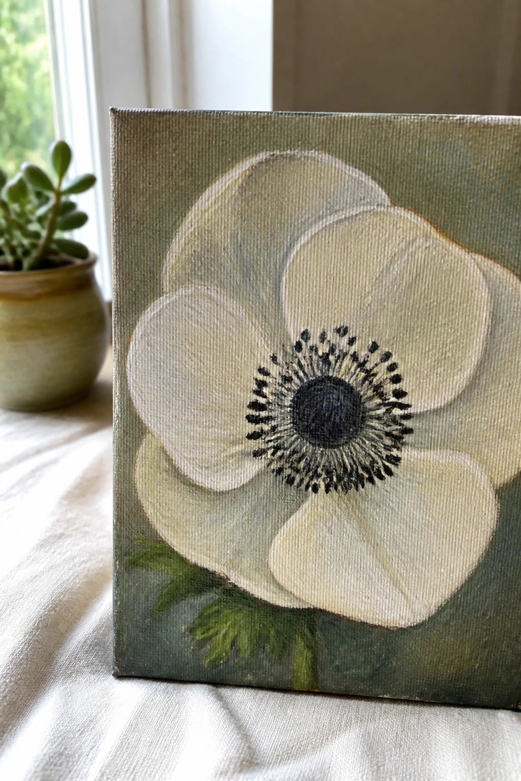 White flower painting with black center on green canvas beside potted plant