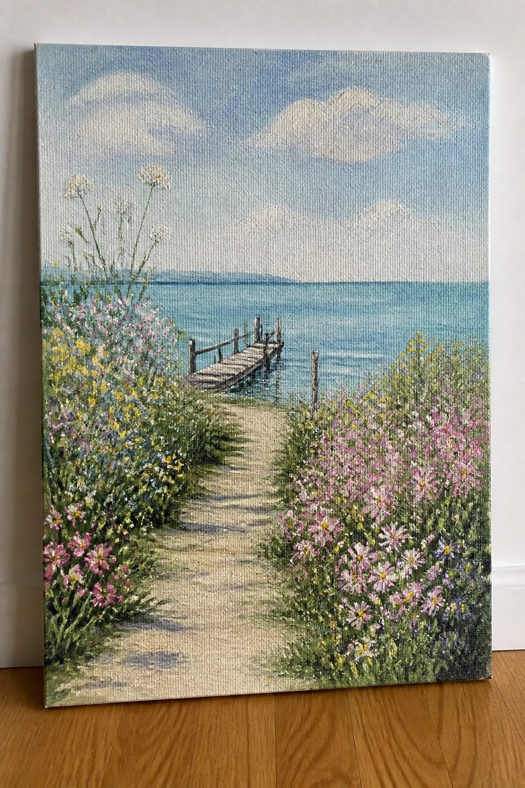 Wildflowers line a sandy path to a wooden dock on calm blue sea under clouds