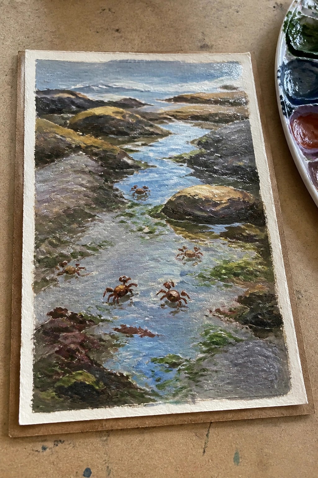 An oil painting of a blue stream flowing through rocky coastal hills with several small brown crabs visible in the shallow water and a paint palette on the right side.