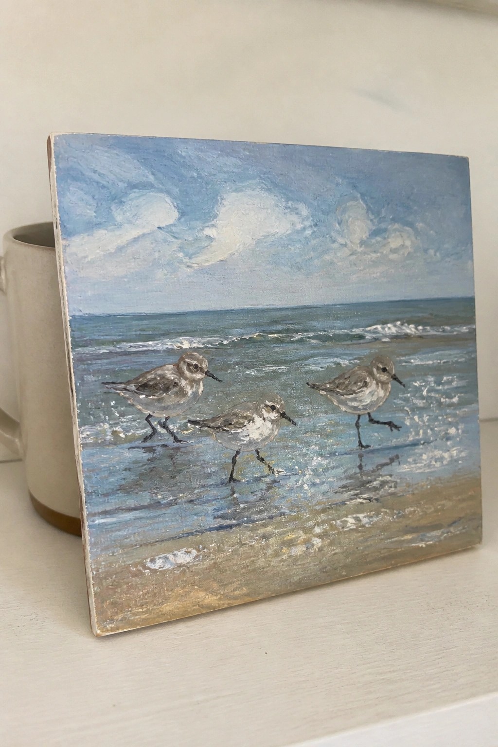 Three sandpipers wade on wet sand in a beach painting beside a beige mug.