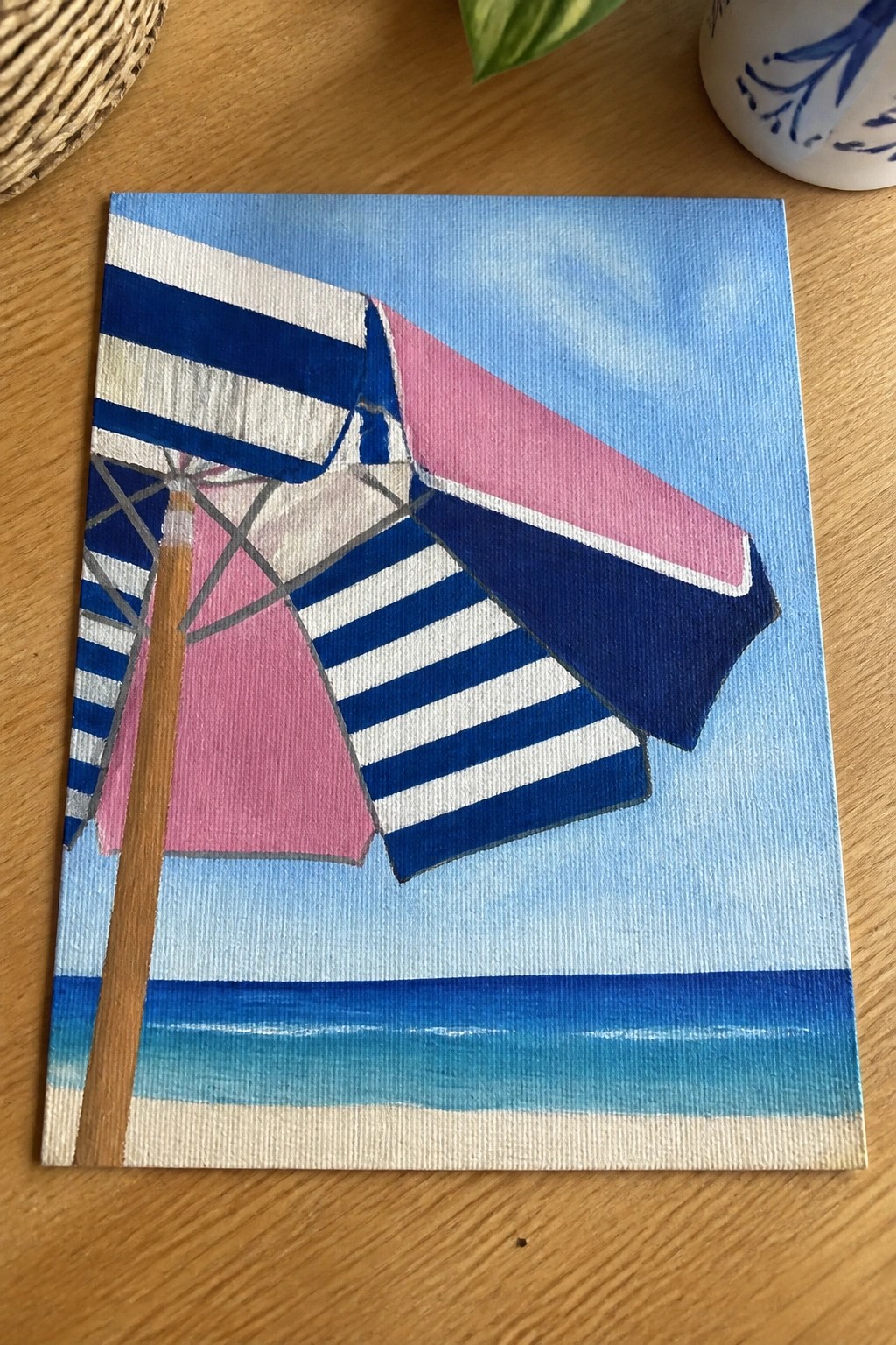 Striped beach umbrella painting in blue, white, and pink over ocean scene