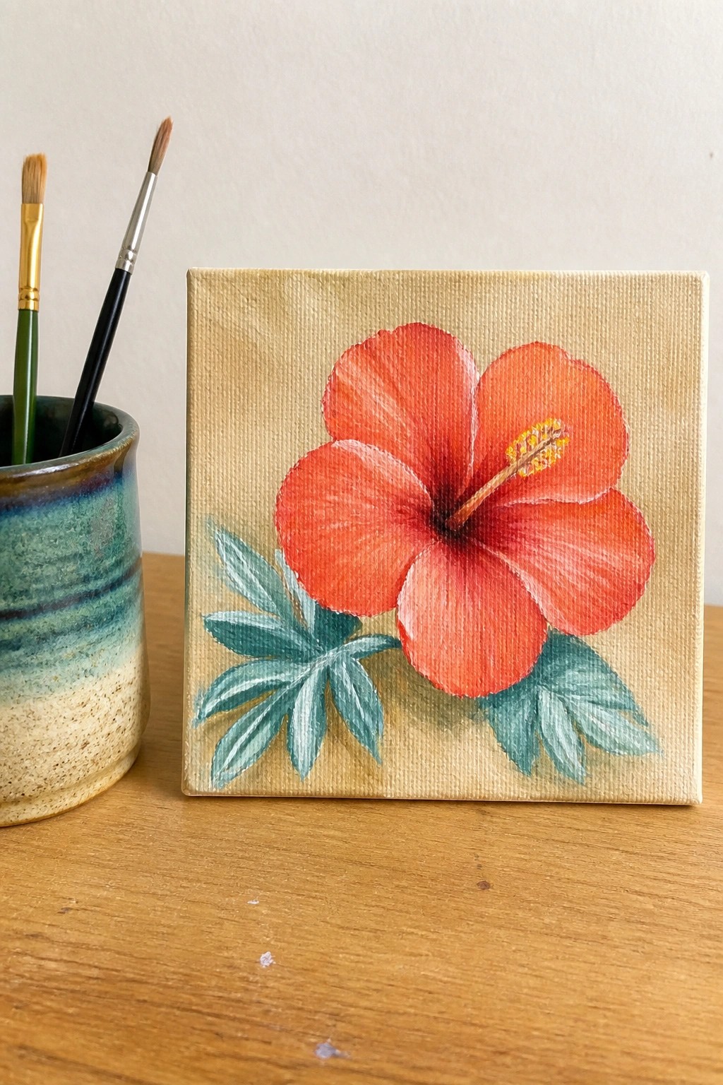 Red hibiscus flower painting on small canvas with paintbrushes nearby.