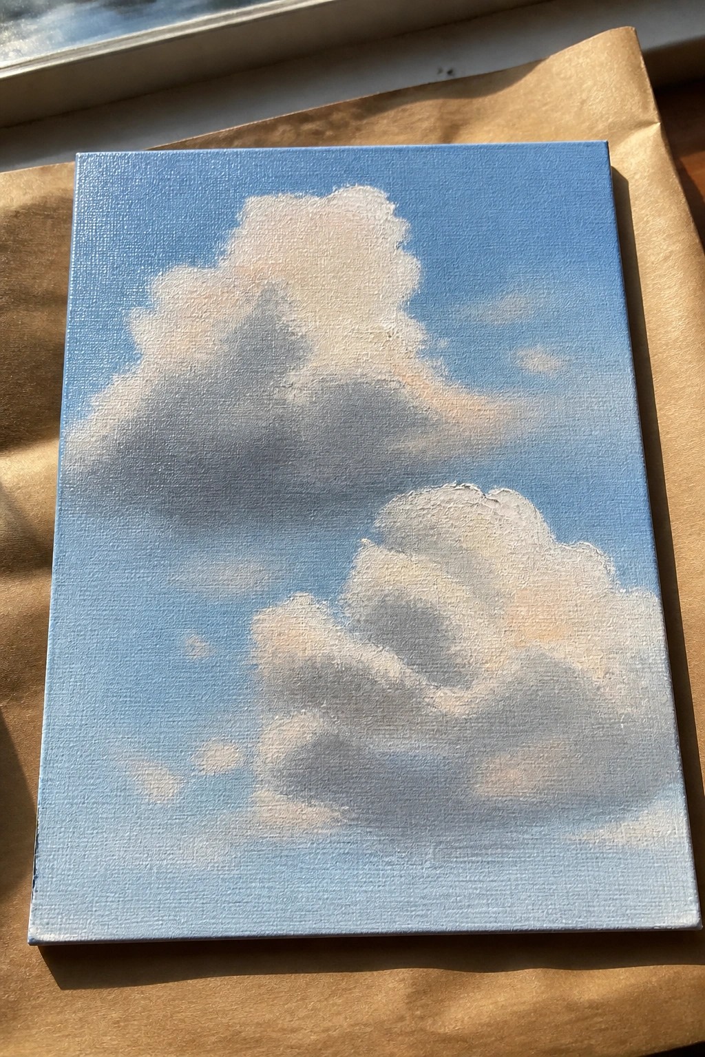 Small canvas painting of fluffy clouds in light blue sky on brown paper