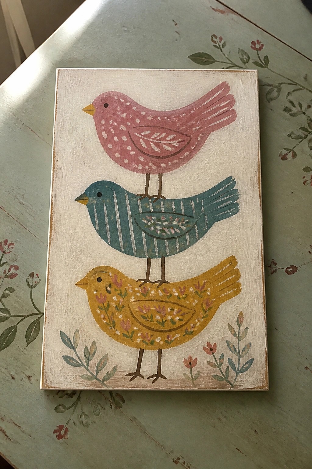 Folk art painting of three stacked birds in pink, teal, and yellow on wood
