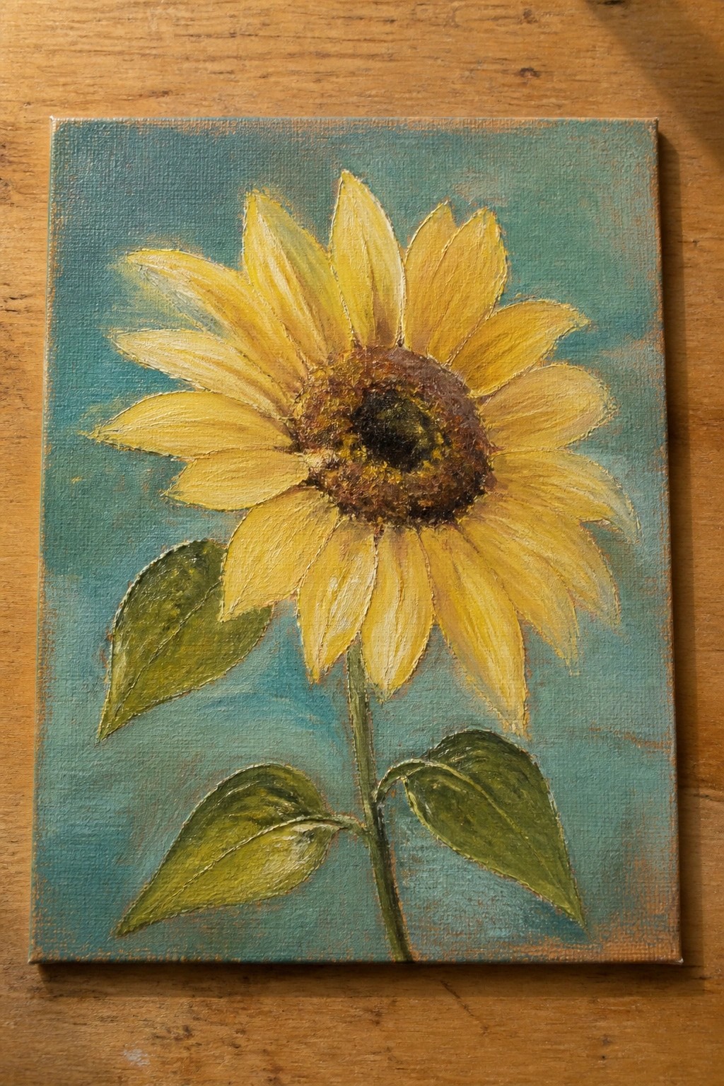 Yellow sunflower painting with green leaves on textured teal canvas over wood