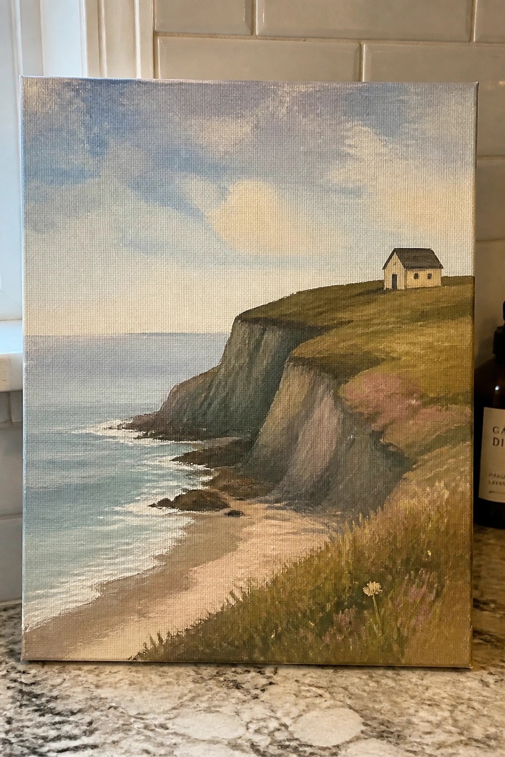 Painting of a house on grassy cliffs above a sandy beach and calm blue ocean.