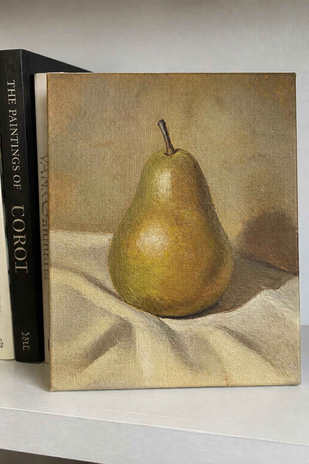 Painting of a green pear on white cloth propped against books on shelf