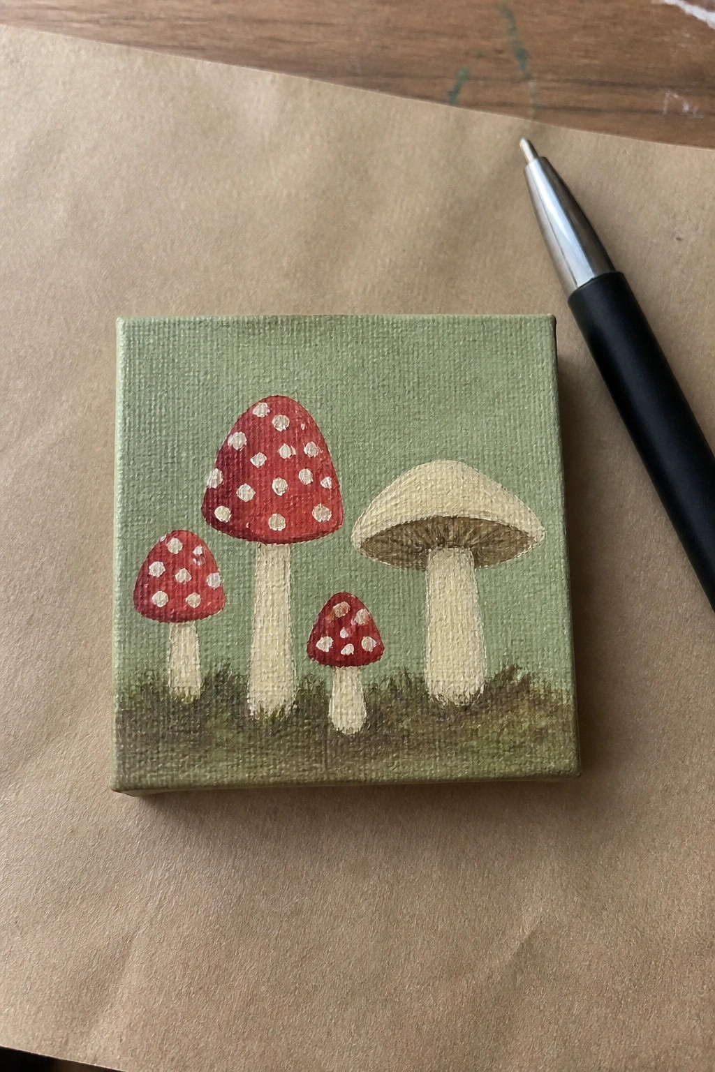 A small square oil painting shows four mushrooms with red spotted and beige caps on a green background next to a black pen.