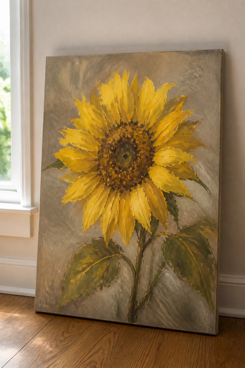 An oil painting of a large yellow sunflower with green leaves against a textured gray background, leaning against a wall on a wooden floor.