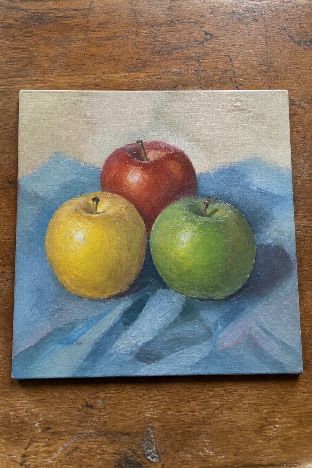 Still life oil painting of yellow, red, and green apples on blue cloth.