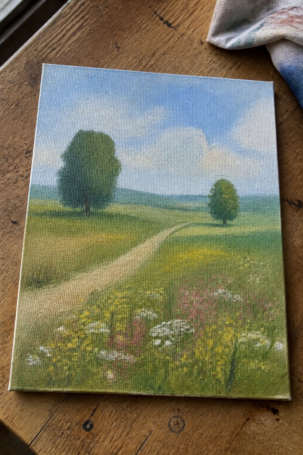 Landscape painting on canvas of path through green fields with wildflowers and trees.